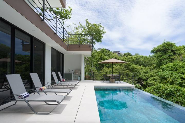 Pool deck with four loungers, the outdoor kitchen and a shaded table over the trees